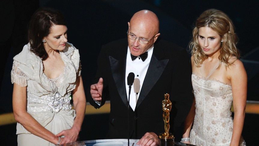 Heath Ledger's mother, father and sister stand at a podium with an Oscar award.