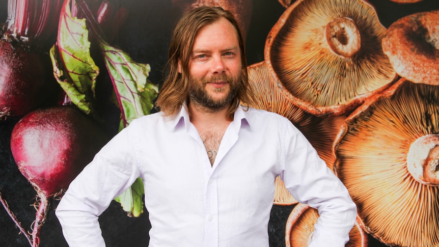 A man with a beard and shoulder-length hair standing in front of a wall photo of vegetables.