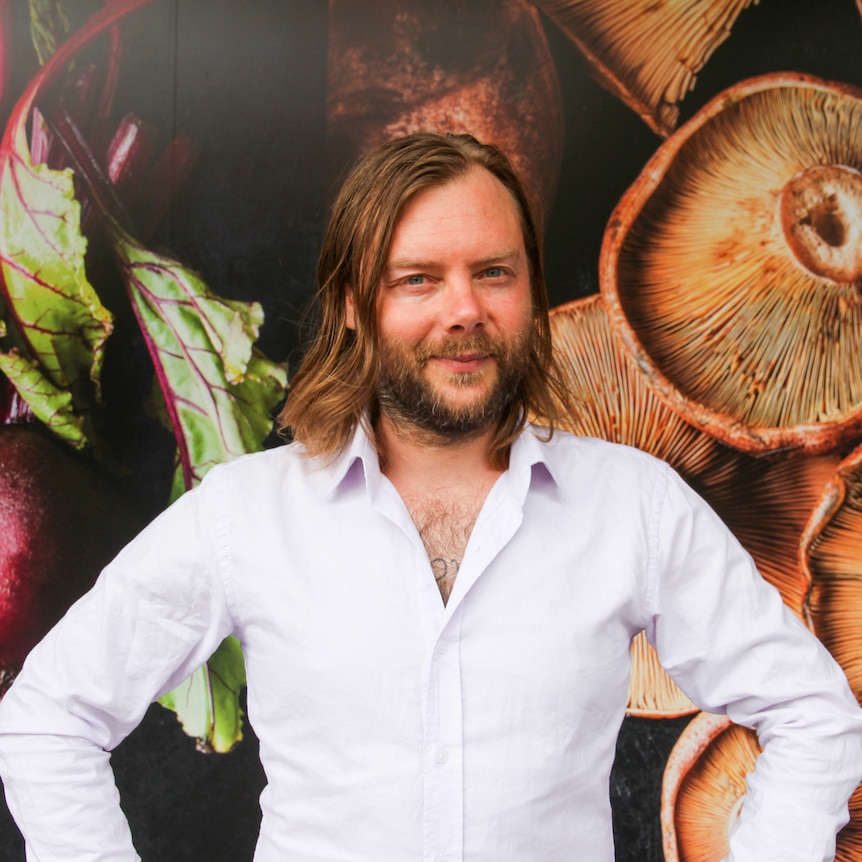 A man with a beard and shoulder-length hair standing in front of a wall photo of vegetables.
