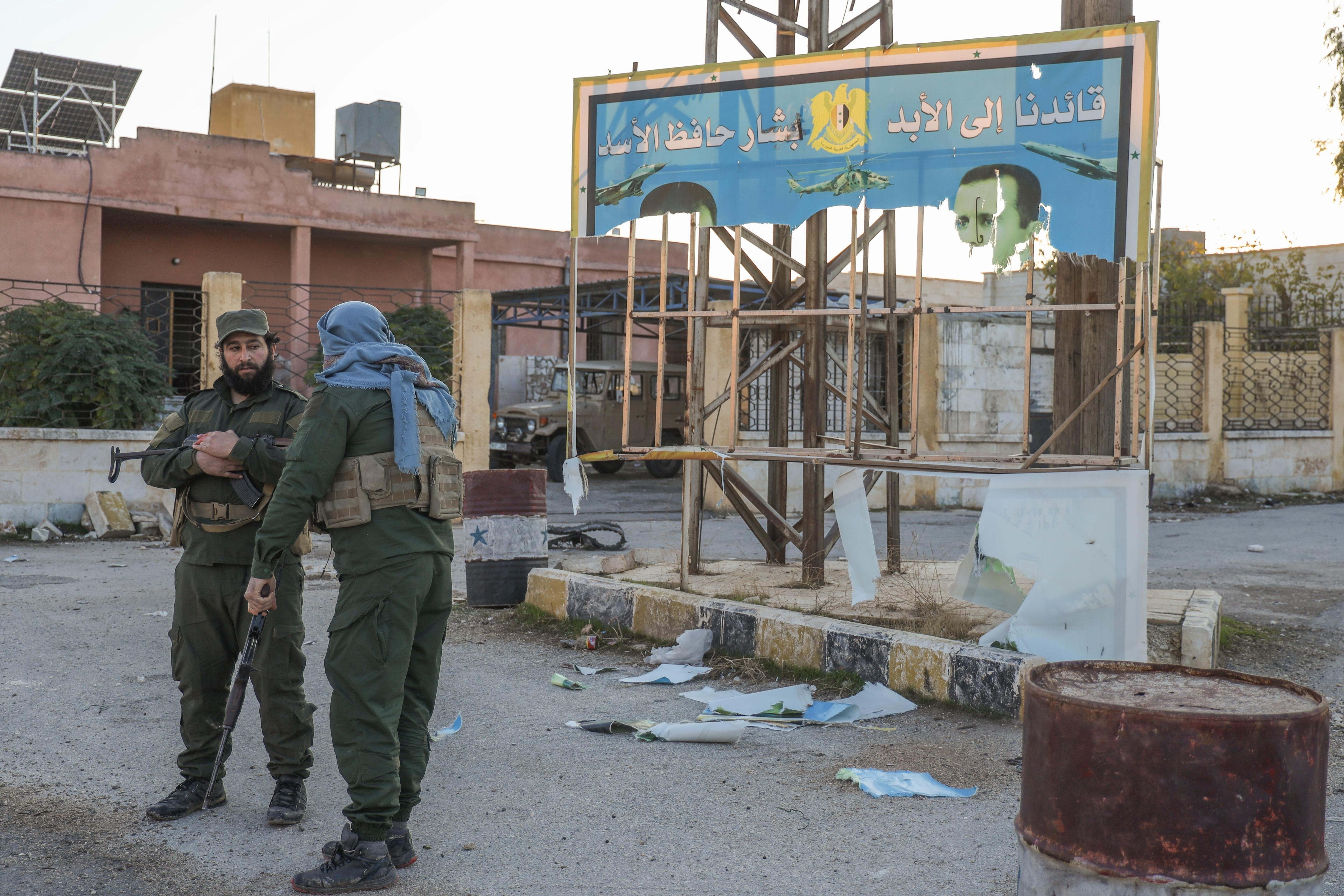 Two men in combat gear stand in front of a wrecked blue billboard.