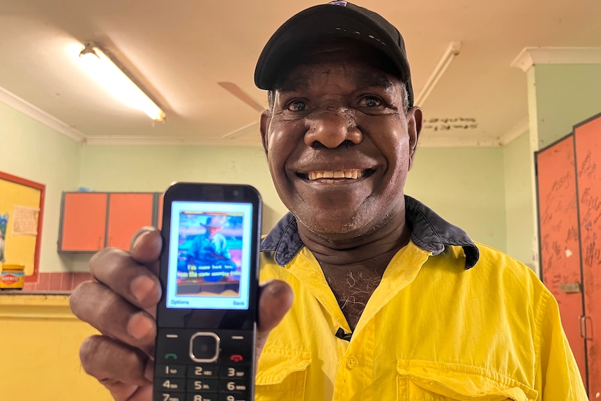 A smiling man wearing a yellow work shirt holds up a mobile phone with a photo of an older man on the screen