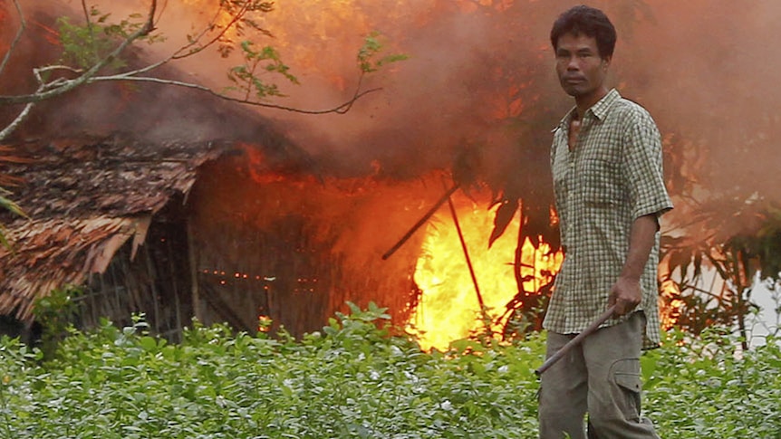 An ethnic Rakhine man holds homemade weapons as he stands in front of a burning house