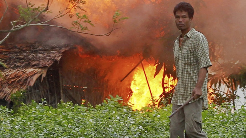 An ethnic Rakhine man holds homemade weapons as he stands in front of a burning house.