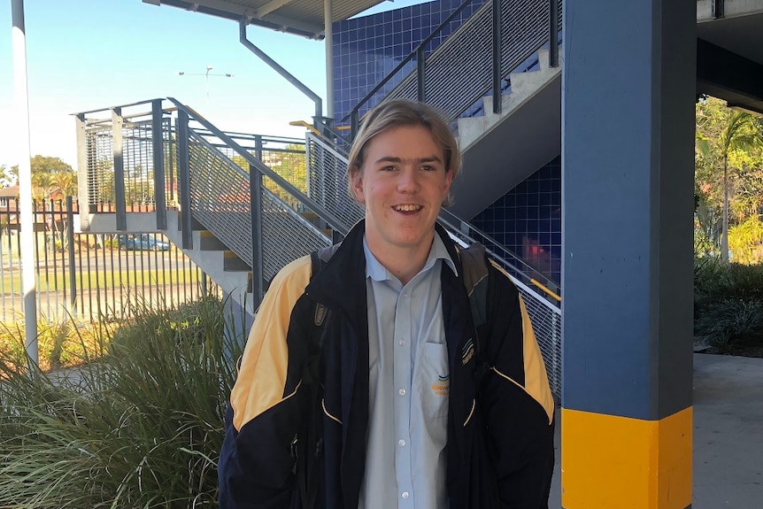 A smiling teenage boy standing in school uniform near stairs of a school building