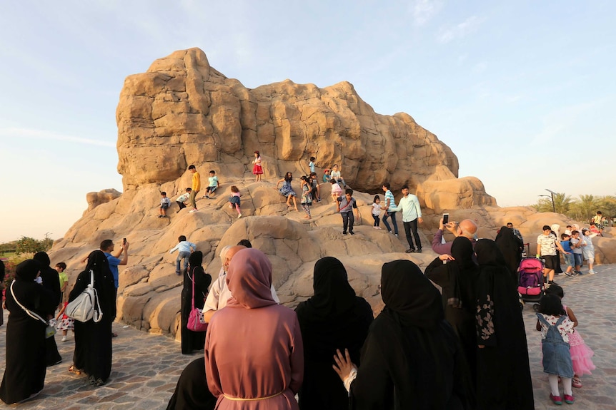 A fake mountain-like rock is seen behind a string of women in black hijabs watching on as their children climb the rock.