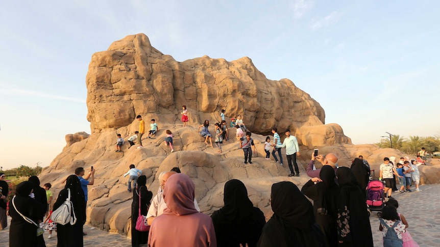 A fake mountain-like rock is seen behind a string of women in black hijabs watching on as their children climb the rock.