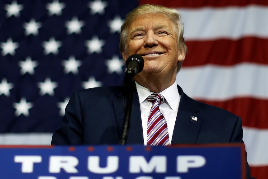 Donald Trump smiles in front of an American flag.