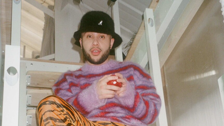 jax jones sitting on awhite wooden lader wearing a bring red and purple jumper, tiger striped pants and a black hat