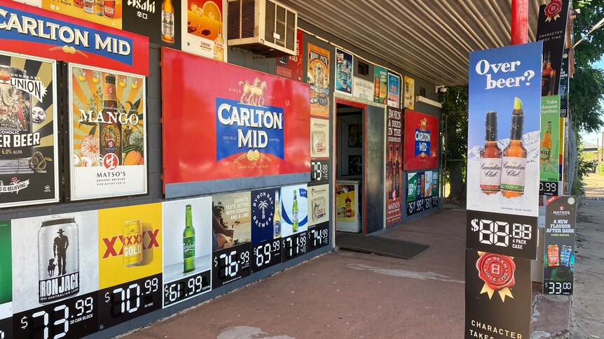 A bottleshop with posters of different drinks, including Carlton, and aircon on the wall under a verandah with cream roof.