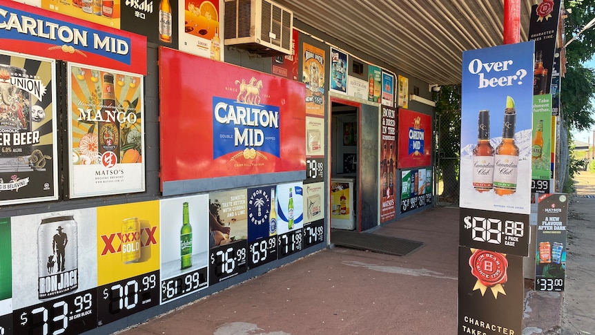 A bottleshop with posters of different drinks, including Carlton, and aircon on the wall under a verandah with cream roof.