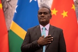 A man in a suit rests his hand on his chest, standing in front of Solomon Island and China flags.