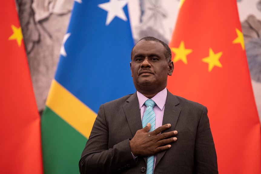 A man in a suit rests his hand on his chest, standing in front of Solomon Island and China flags.