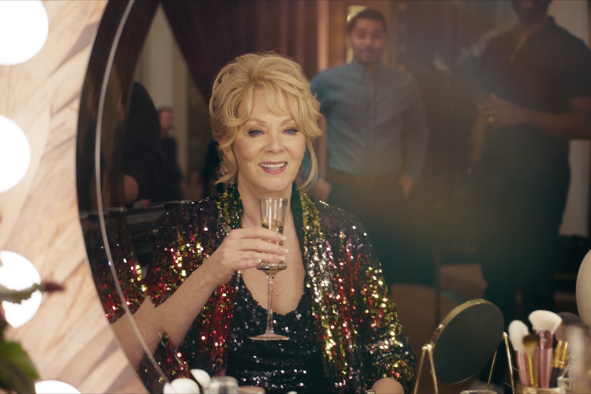 A woman in her late 60s with blonde hair and wearing a sparkly outfit, holds a glass of champagne and looks into a mirror