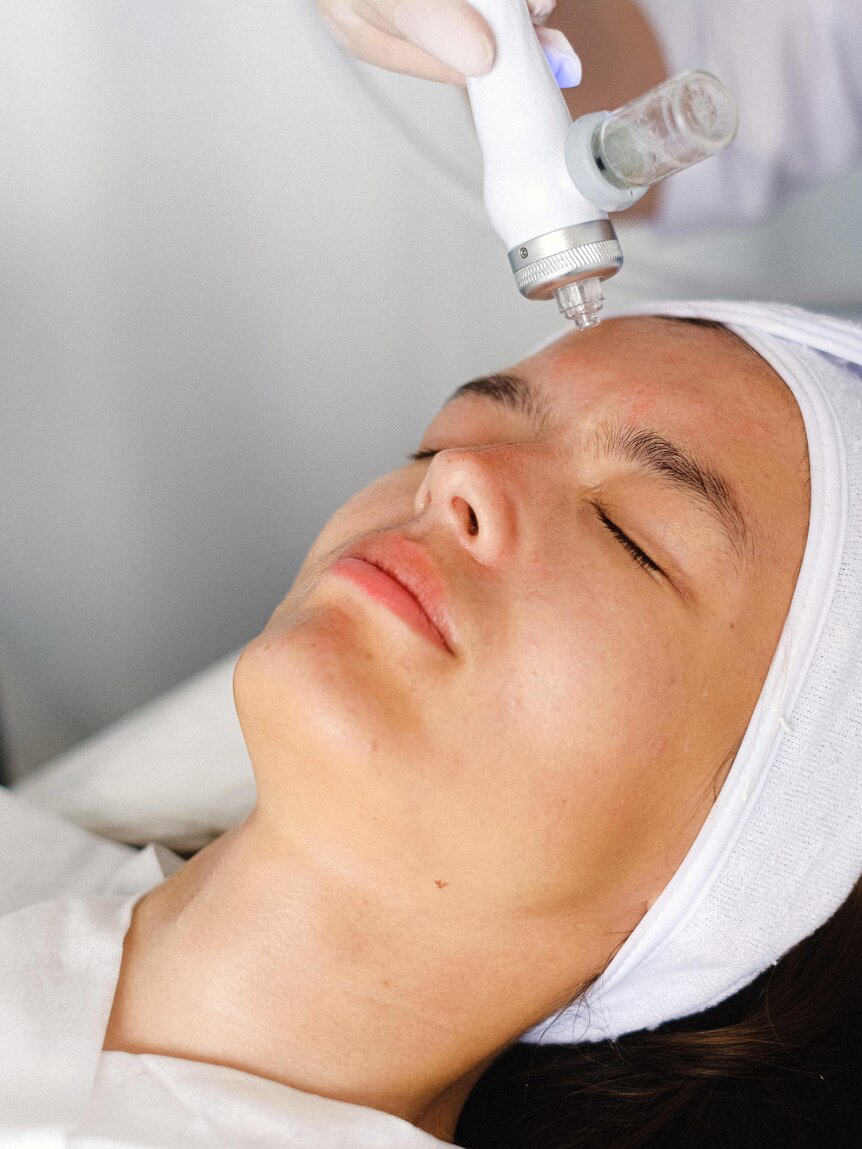 A woman lies in a salon bed with a microdermabrasion tool being used on her face.