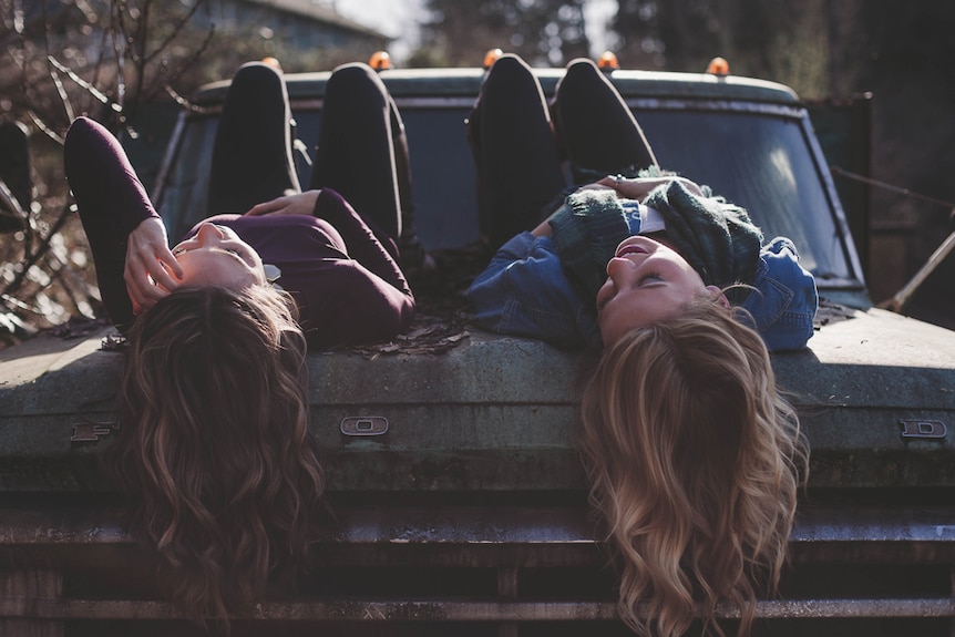 Two women lying on car bonnet and having a chat rather than messaging through social media.
