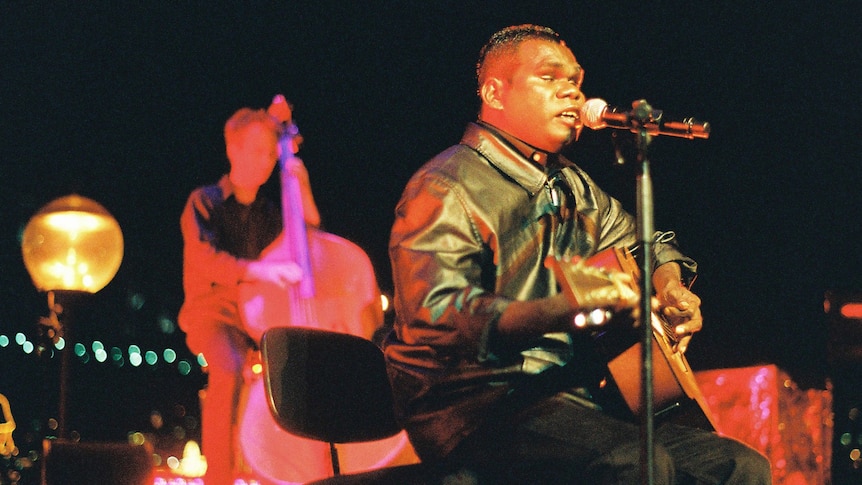 Gurrumul plays guitar and sings onstage. He wears a leather jacket. Behind him stands a double bass player.