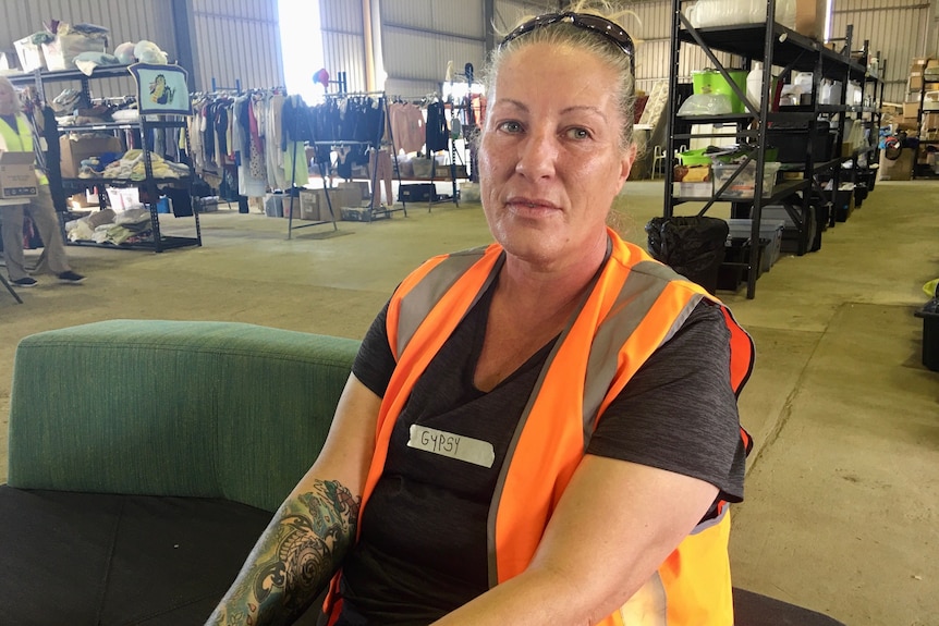 Woman with hair pulled back, wearing black t-shirt and orange hi-vis vest, sitting in a warehouse