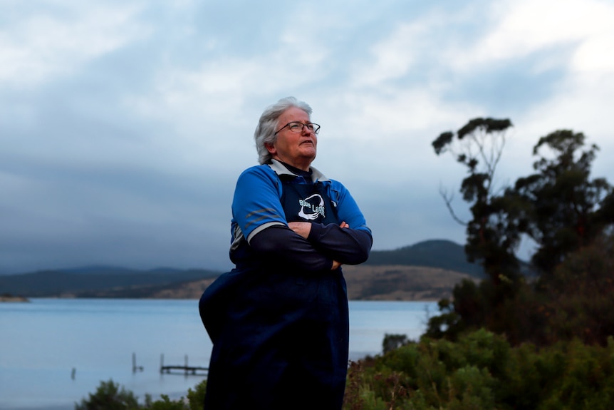 Woman with white hair wearing navy blue apron and blue shirt stands outside with bay and moody coastline behind.