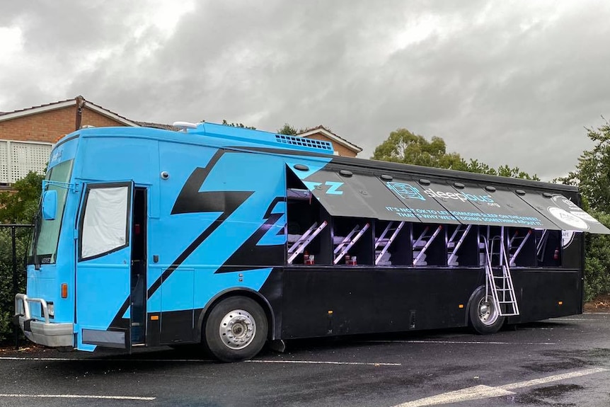 A black and blue painted bus with side doors open containing sleeping pods. 
