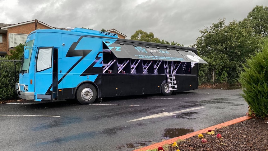 A black and blue painted bus with side doors open containing sleeping pods. 