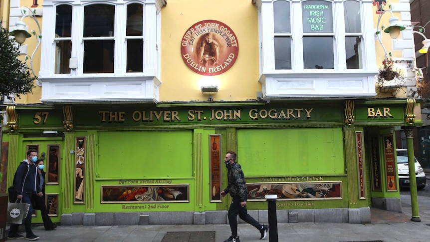 A green pub front shop is boarded up as pedestrians in masks past