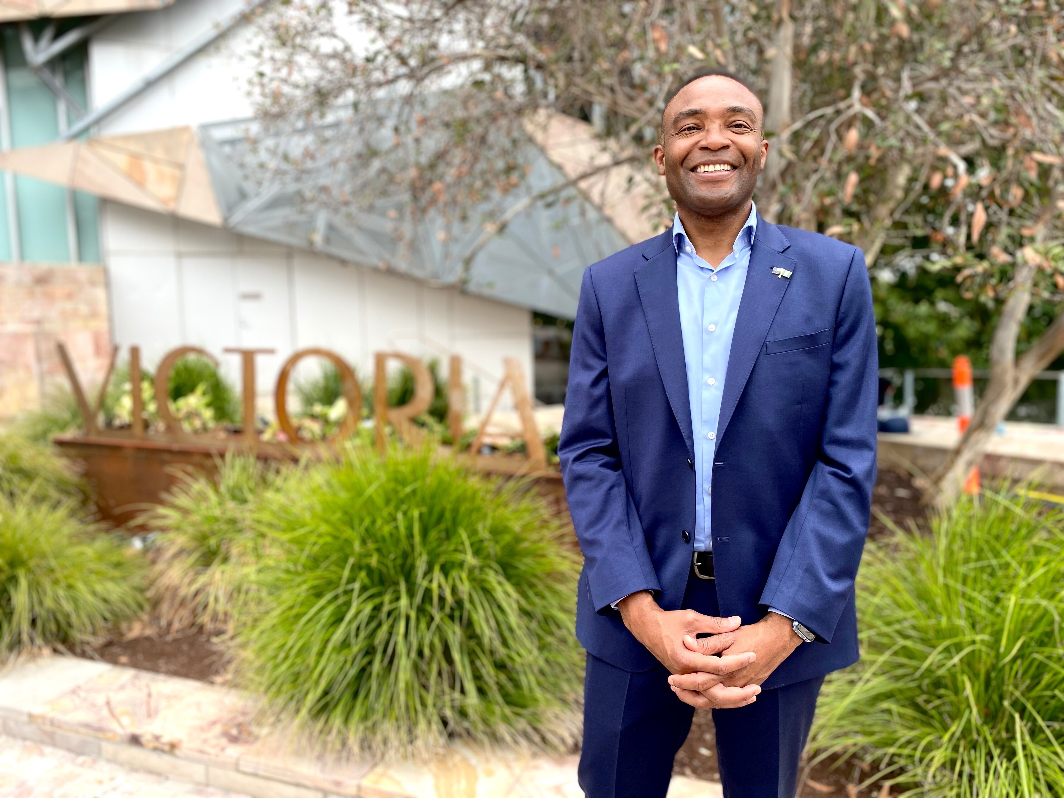 A smiling man in a blue suit stands in front of a sign that says Victoria