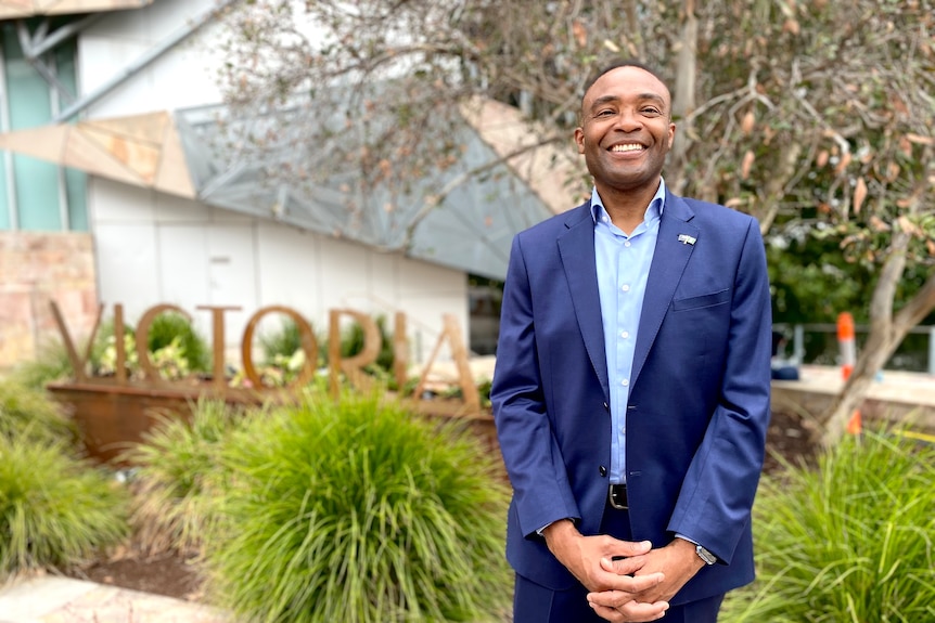 A smiling man in a blue suit stands in front of a sign that says Victoria