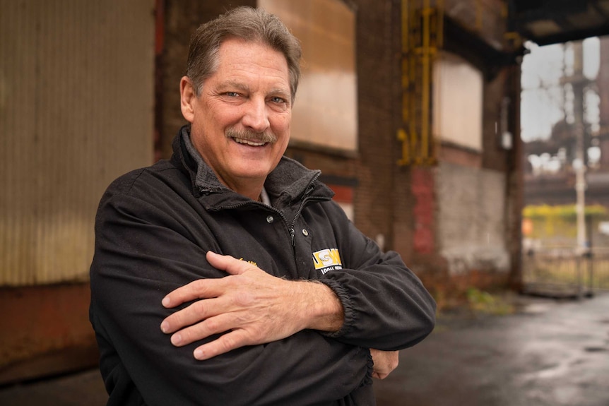 A man smiles with his arms folded over his chest outside an oil refinery