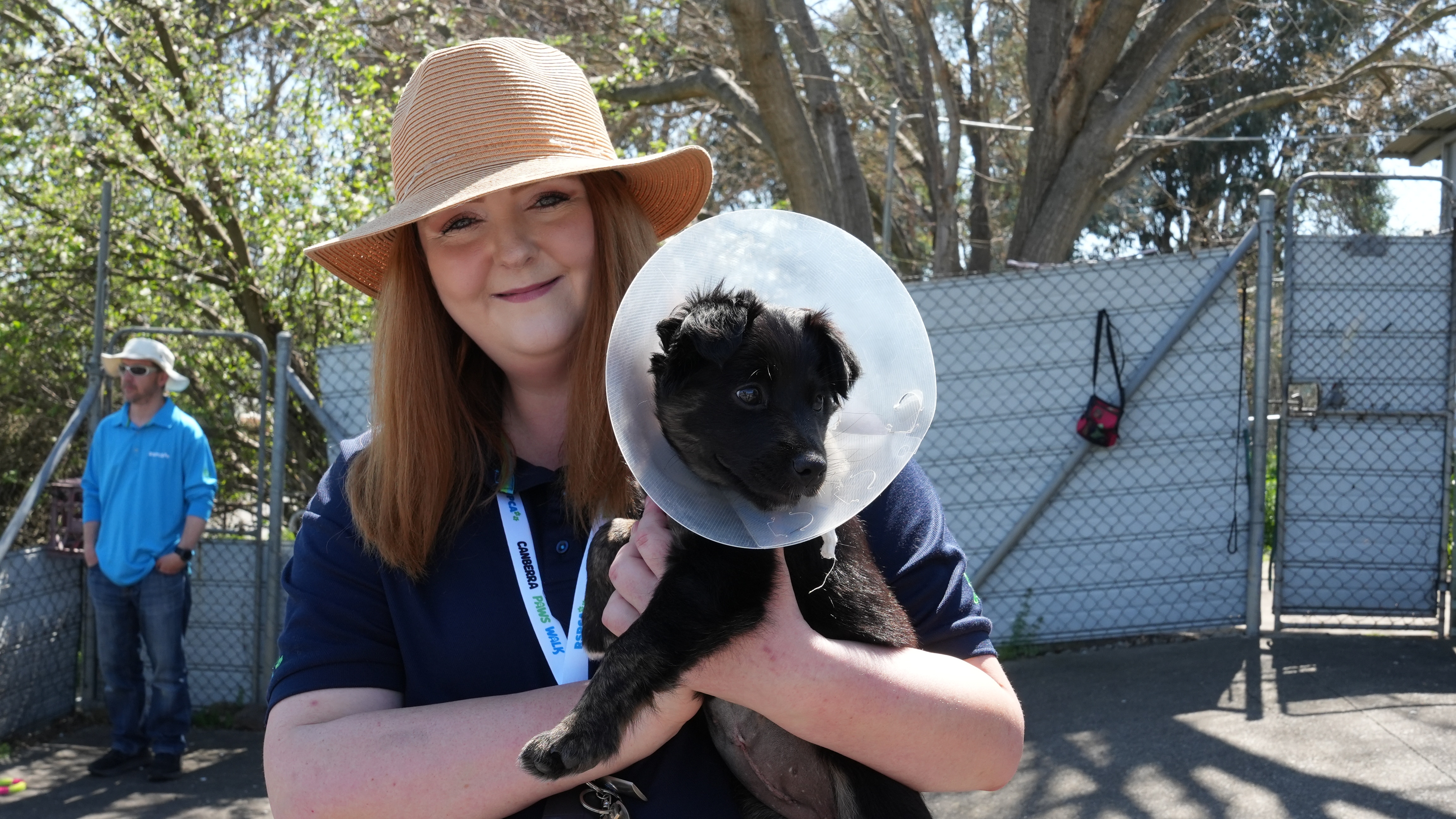 A female RSPCA employee with straight brown hair and a sun hat holds a black puppy with a plastic cone around its head.