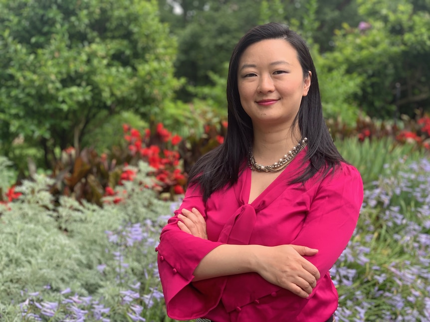 An Asian woman with long black hair wearing a pink jacket and standing in front of flower bed