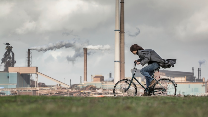 A cyclist riding into a headwind with a factory in the background.