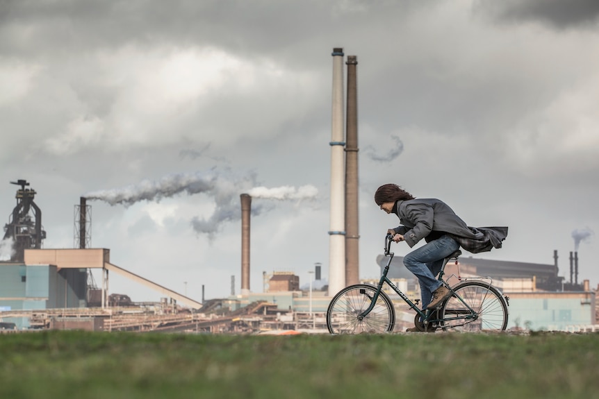 A cyclist riding into a headwind with a factory in the background.