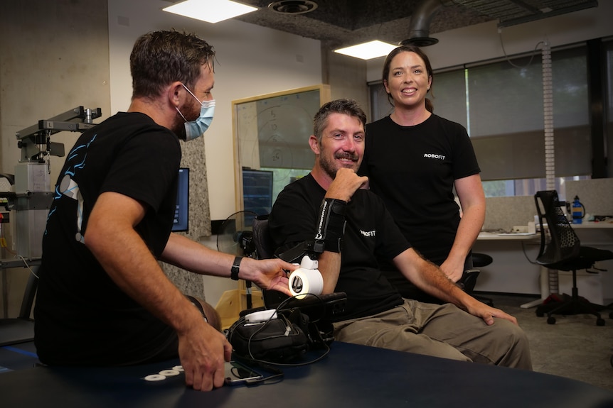 Daniel Hillyer moves his left arm while wearing a robotic attachment, with wife Maryanne standing next to him.