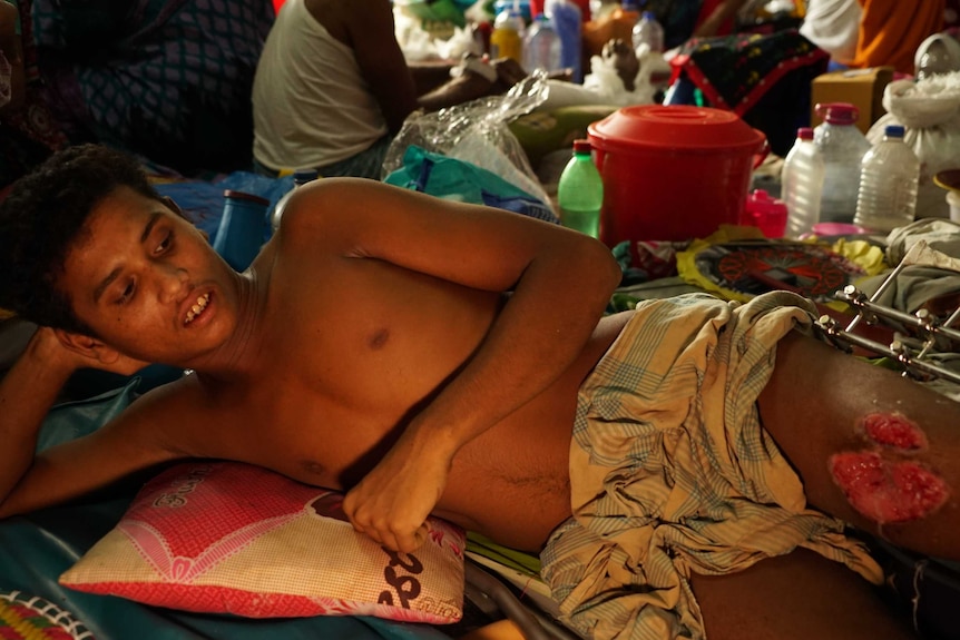 A young man lies on a hospital floor with a gaping wound on his leg.