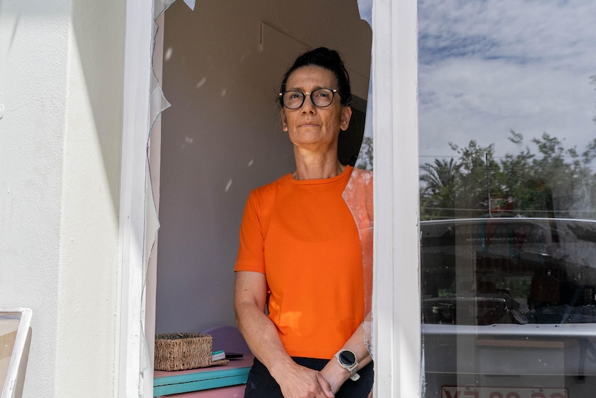 A woman wearing an orange shirt stands on the inside of her home looking out a broken window