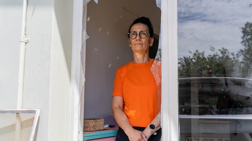 A woman wearing an orange shirt stands on the inside of her home looking out a broken window