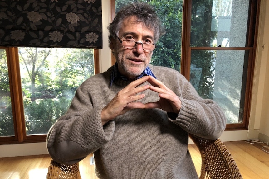 A man with greying hair and glasses sits in a chair and looks thoughtfully at the camera.