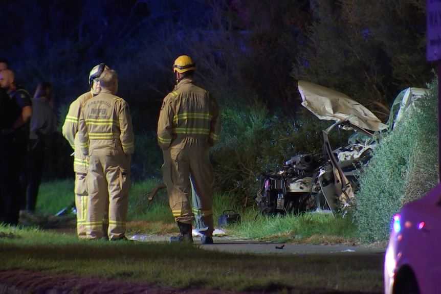 Emergency services workers stand on a road at night, with a mangled car involved in a crash beyond them.