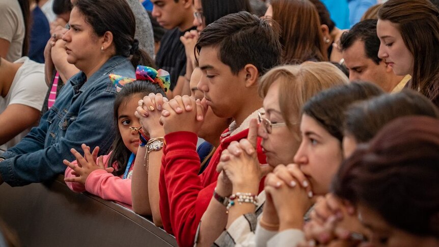 A group of people of various ages pray in a church with their hands clasped and heads bowed