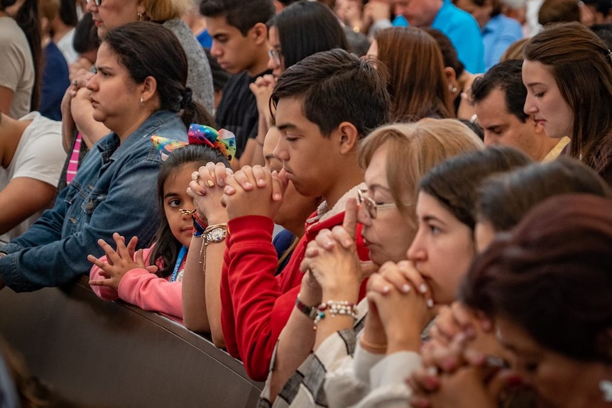 A group of people of various ages pray in a church with their hands clasped and heads bowed