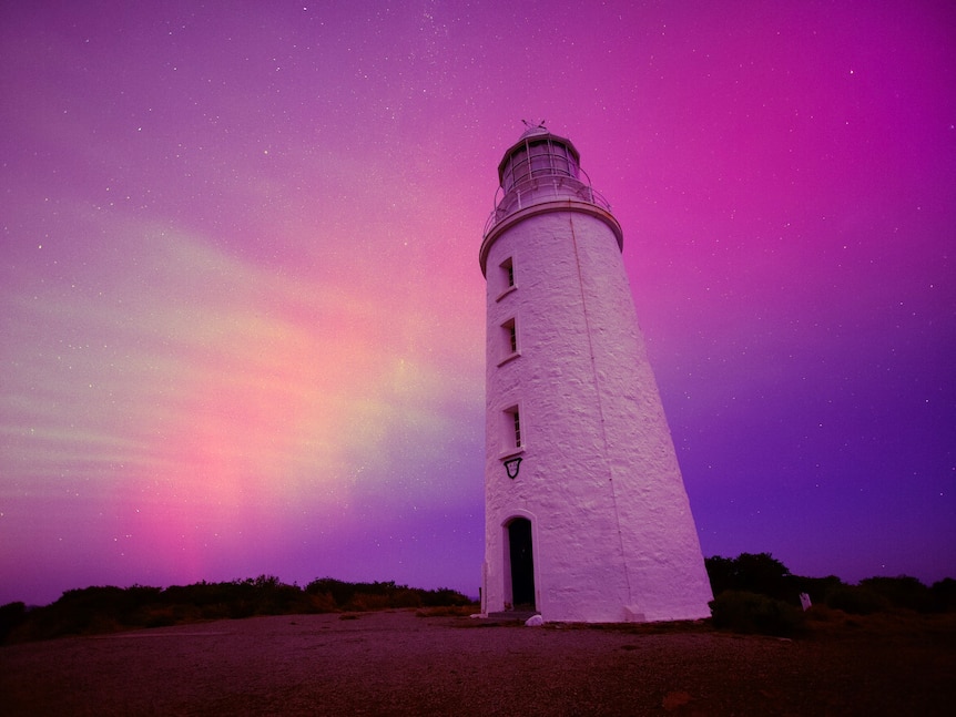 A lighthouse stands in front of a pink and purple sky lit by aurora