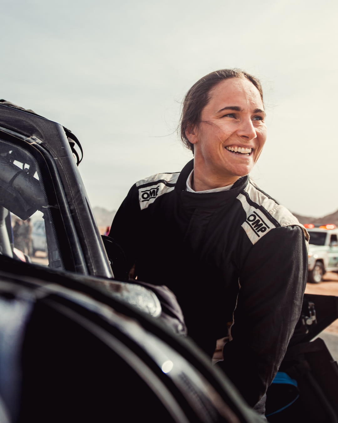 A smiling female rally driver stands next to a vehicle looking off in the distance away from the camera.