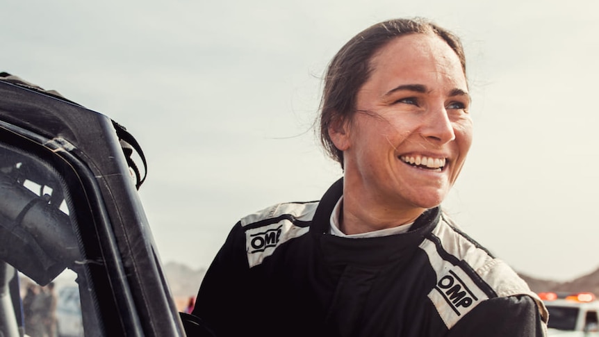 A smiling female rally driver stands next to a vehicle looking off in the distance away from the camera.