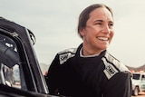 A smiling female rally driver stands next to a vehicle looking off in the distance away from the camera.
