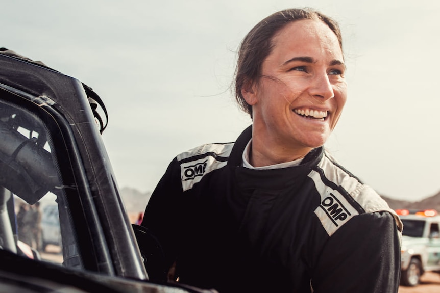 A smiling female rally driver stands next to a vehicle looking off in the distance away from the camera.