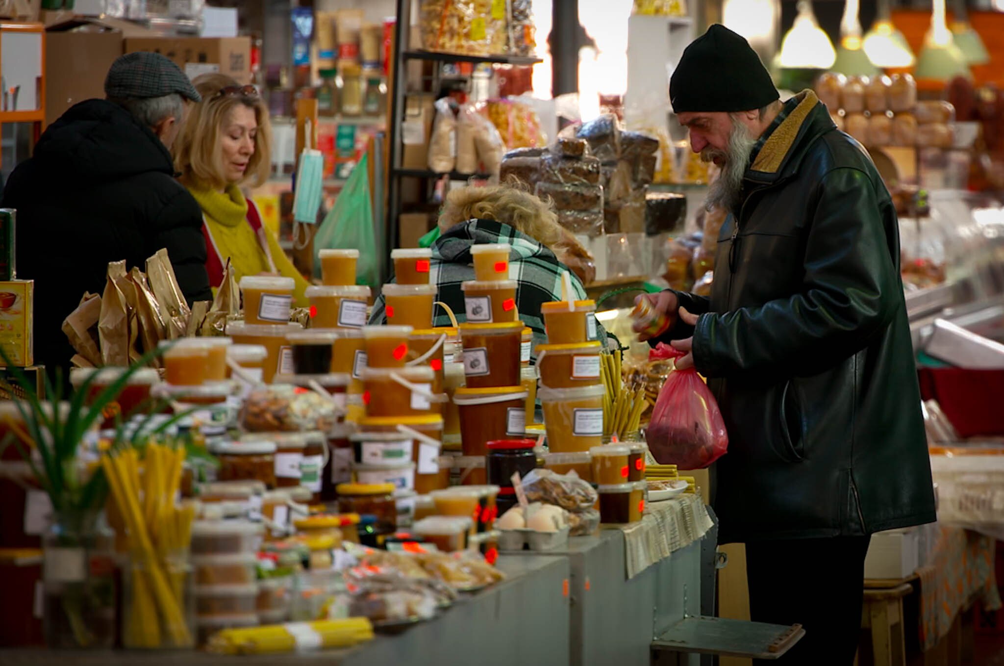 a bearded man peruses pots of honey on a tressel table in a market