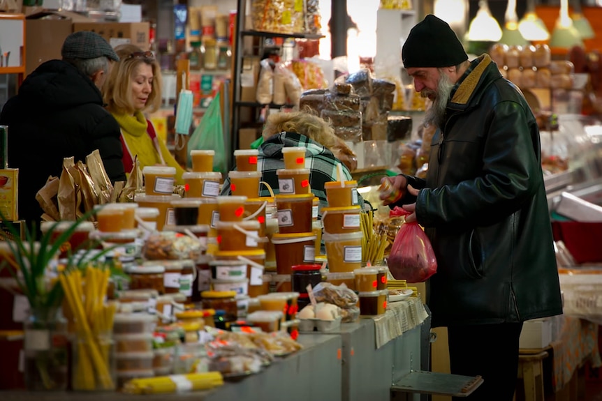 a bearded man peruses pots of honey on a tressel table in a market