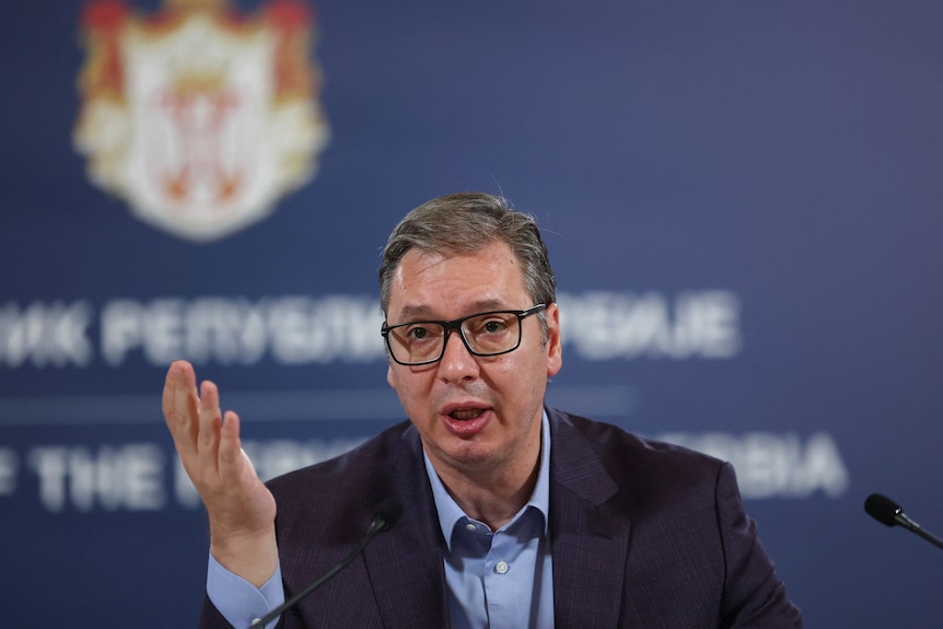 A middle-aged white man with glasses in a suit gestures as he speaks into a microphone in front of a blue backdrop.