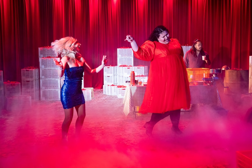 A middle-aged woman in a blonde wig and a full-figured 20-something in a red dress dance on stage, an Asian man standing behind 