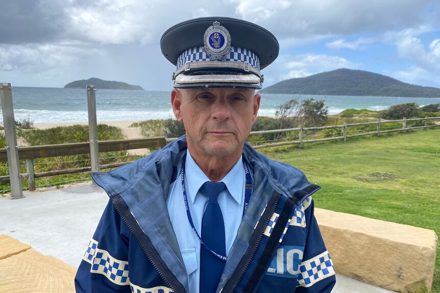 Policeman in uniform looks at camera with serious expression, with beach behind him.